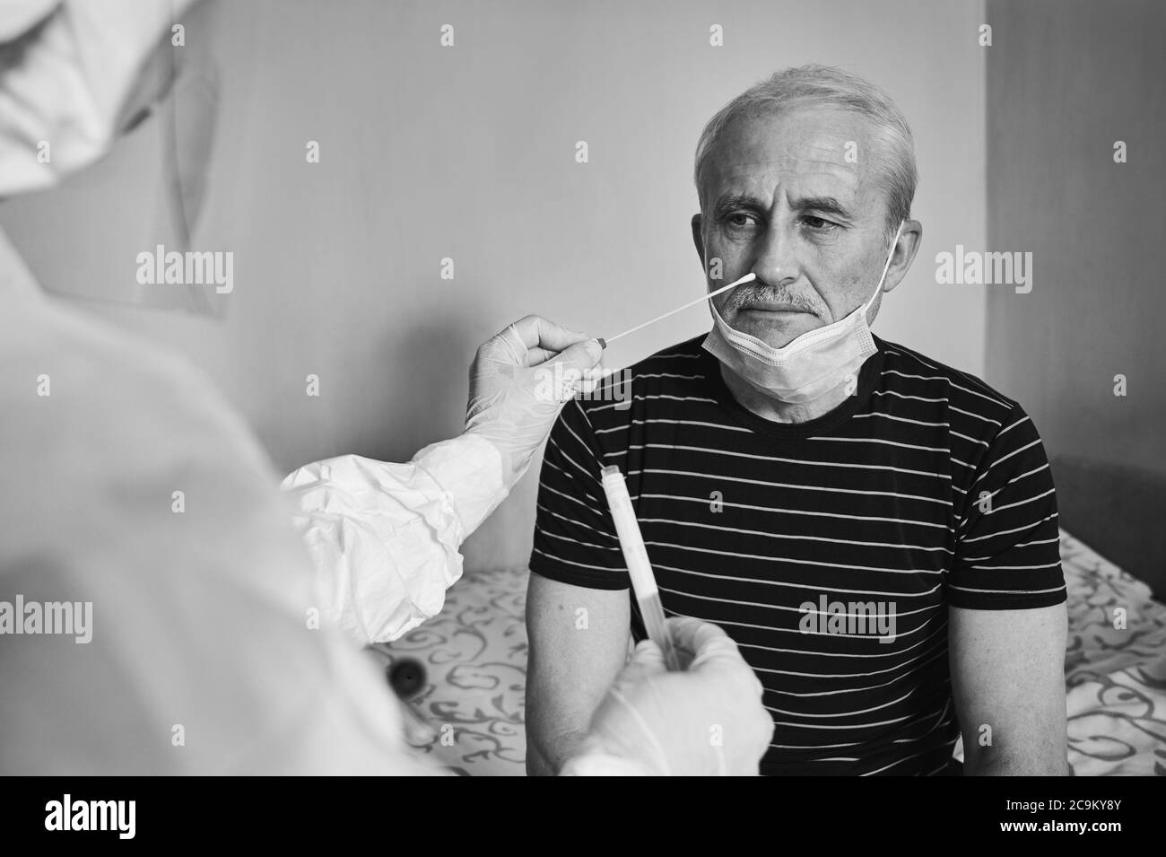 Black and white snapshot of doctor's visit at home. Nurse taking coronavirus test analysis with medical swab to elderly male patient. Test tube for taking OP NP patient specimen sample. Stock Photo