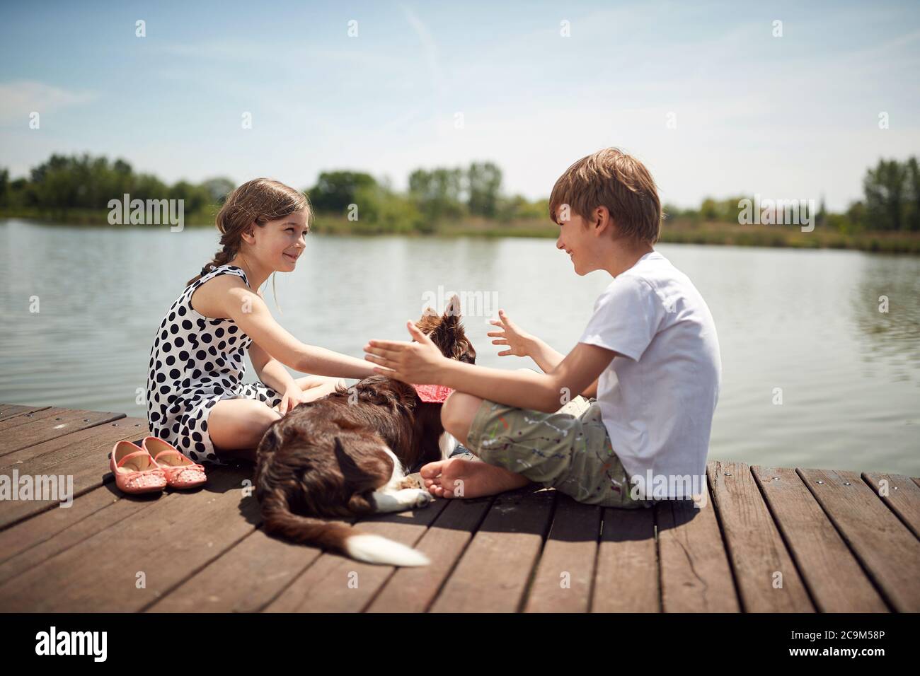 Brother and sister enjoying on the dock of the lake with their dog Stock Photo