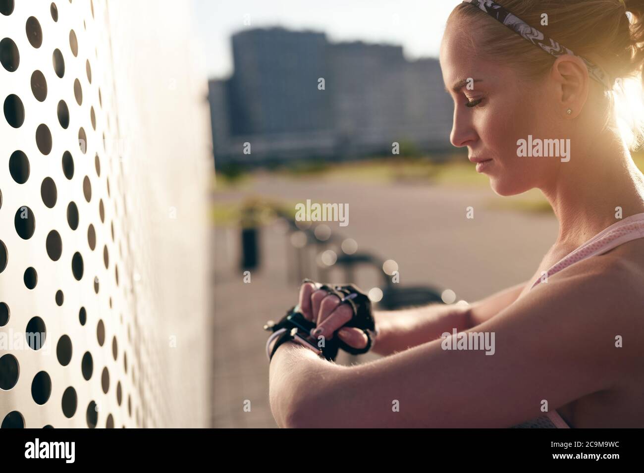 Fitness muscular woman is checking fitness tracker. Sporty girl is using smart watch to track weight in fitness app. Back lit, city view Stock Photo