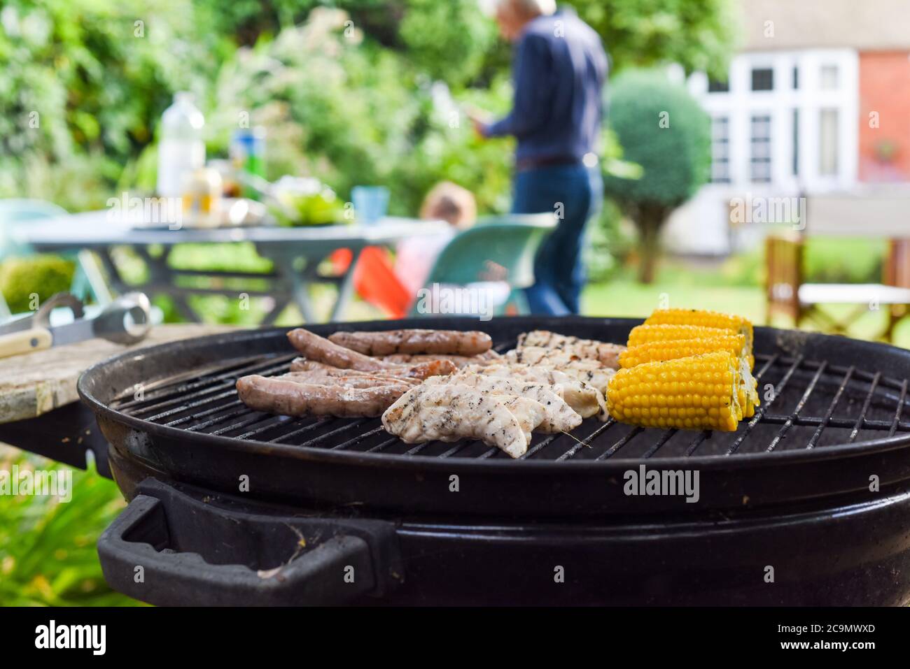Family bbq outside in a garden with food on the barbecue grill cooking outdoors and people in the background Stock Photo