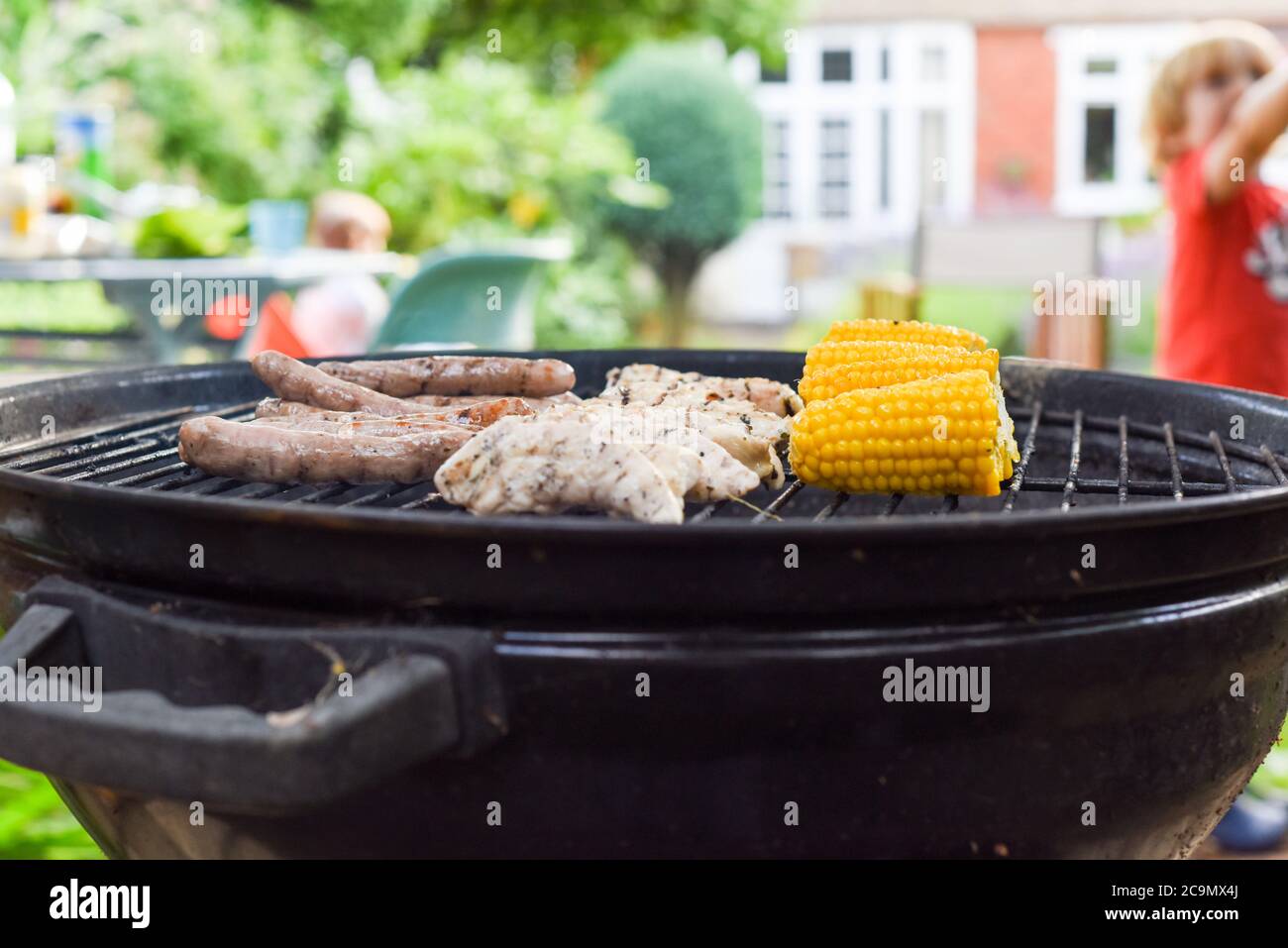 Family bbq outside in a garden with food on the barbecue grill cooking outdoors and people in the background Stock Photo