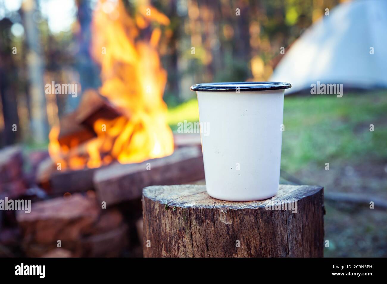 Close up of white camping cup, on top of wood stump, in front of campfire and sleeping tent. Stock Photo