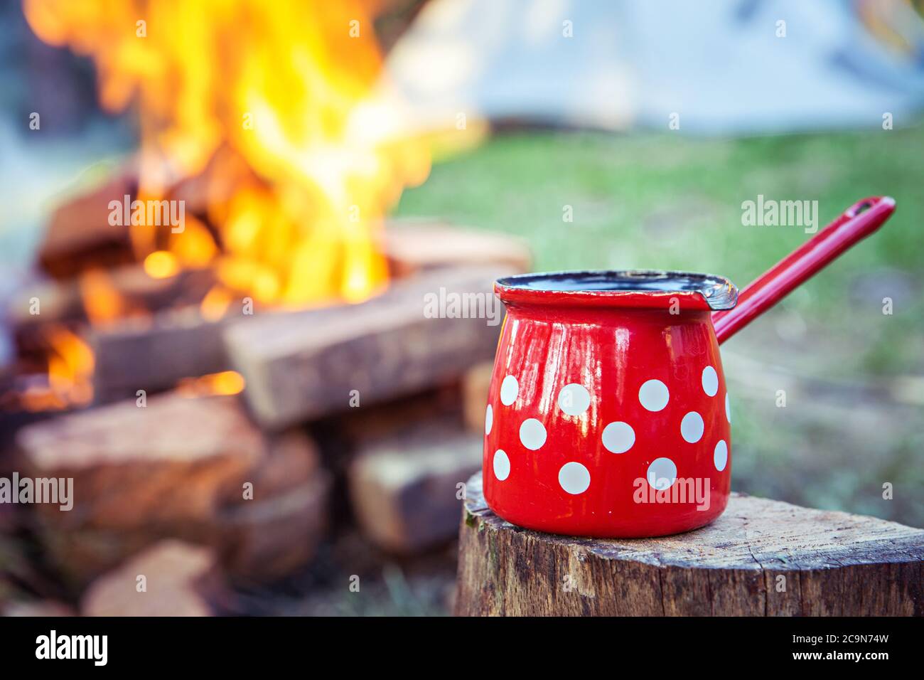 https://c8.alamy.com/comp/2C9N74W/close-up-of-red-coffee-pot-on-top-of-wood-stump-with-campfire-and-camping-tent-in-the-background-2C9N74W.jpg