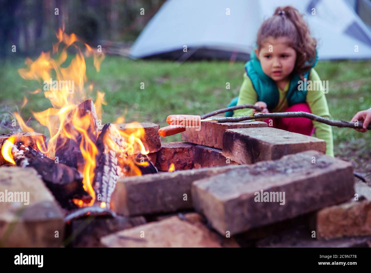 Close up of a young female child, cooking food on a campfire, with camping tent in the background. Stock Photo
