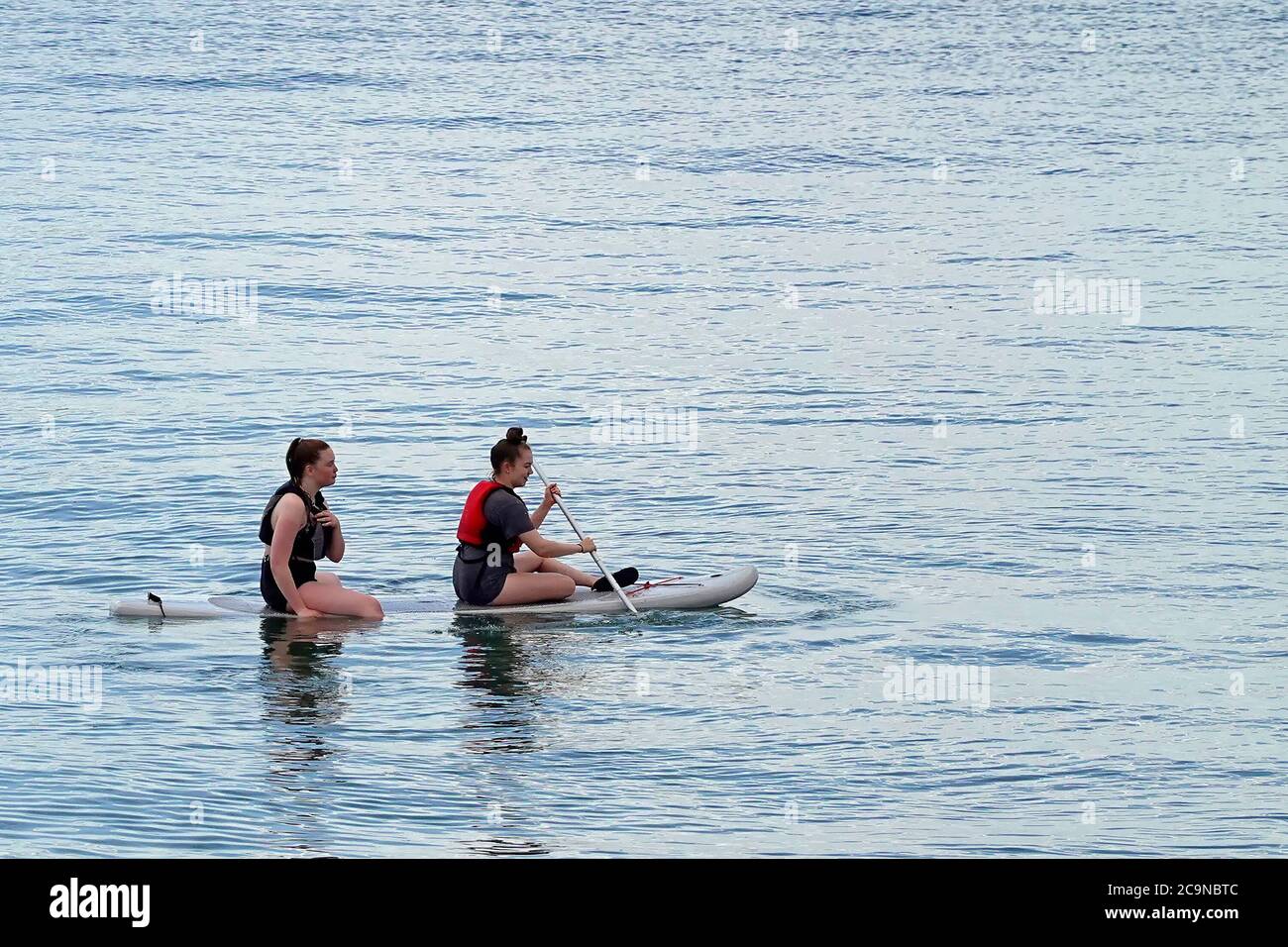 Two women sitting & paddle boarding on paddle board in calm tranquil water on River Thames estuary close to shoreline Southend on Sea Essex England UK Stock Photo
