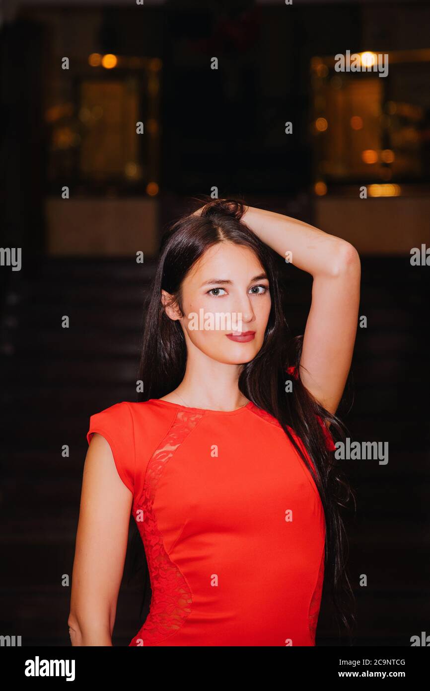 Portrait of a pretty slim female with long dark hair, wearing a stylish long red dress; she has one hand in her hair and looks at us Stock Photo