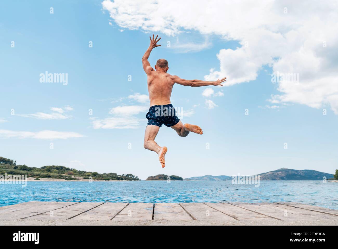 Middle-aged man dressed swimming trunks funny jumping to the waves from the boat pier as he having fun on merry vacation days on Adriatic sea coast. C Stock Photo