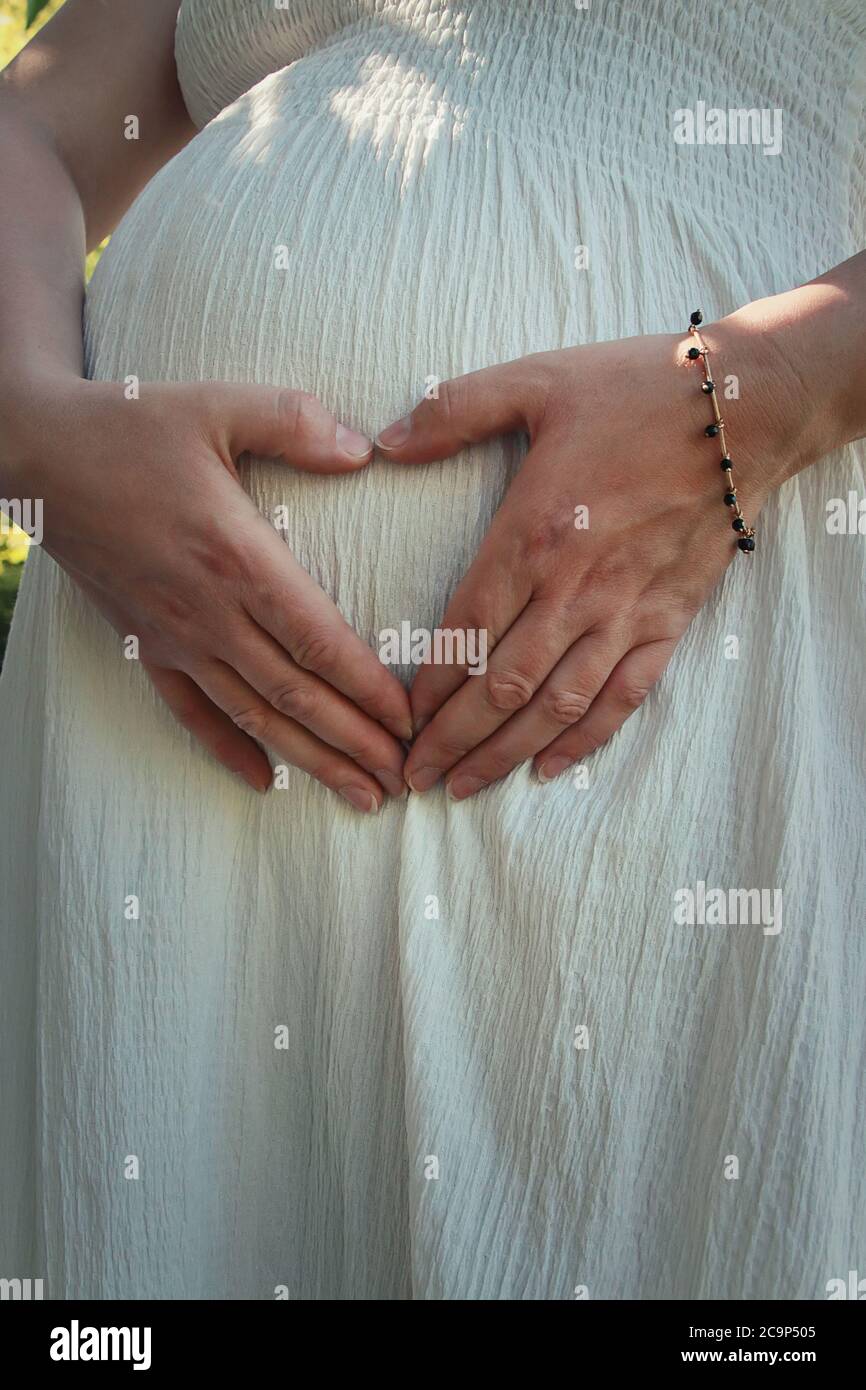 Closeup cute composition with a pregnant young woman wearing white dress makes a heart shaped symbol with her hands on her belly. Happy mother waiting Stock Photo