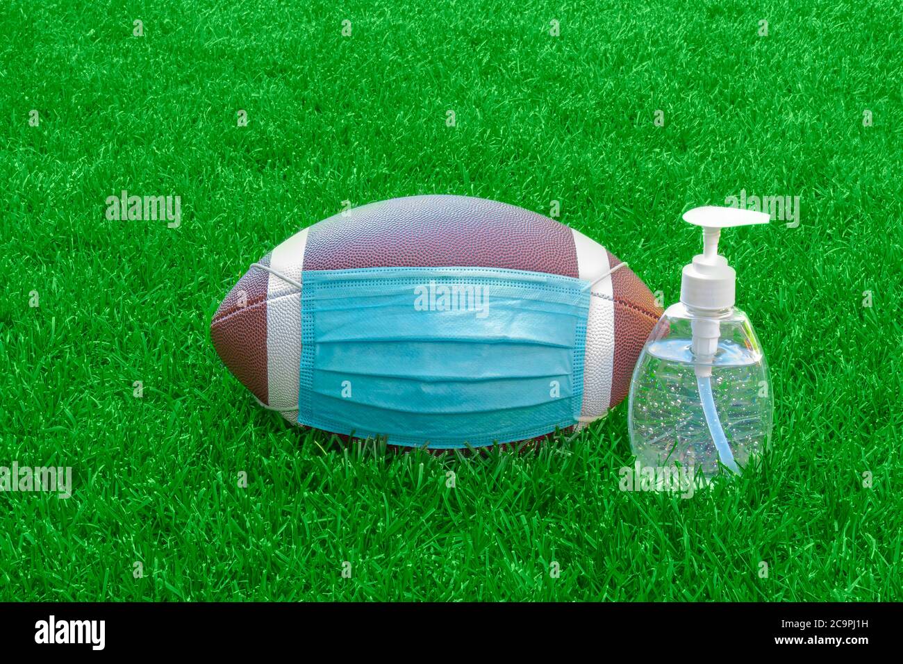 An American Football with a face mask and hand sanitizer on the right on field with green grass. Concept Football during pandemic Stock Photo