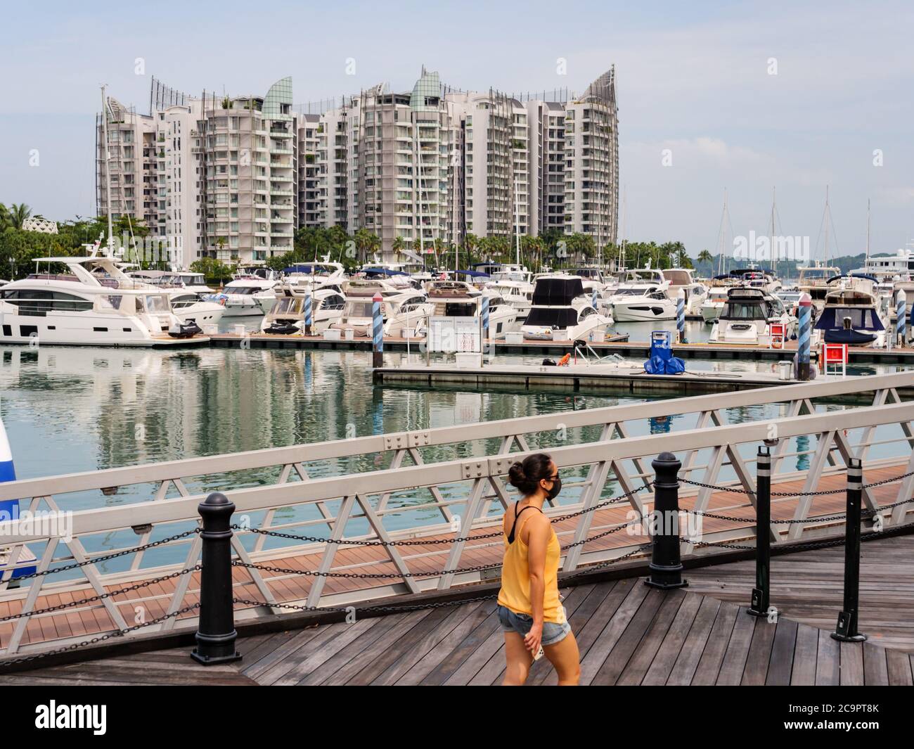 SINGAPORE – JUL 31, 2020 – Asian woman walks past a row of recreational boats docked at Sentosa Island with luxury condominiums in the background Stock Photo