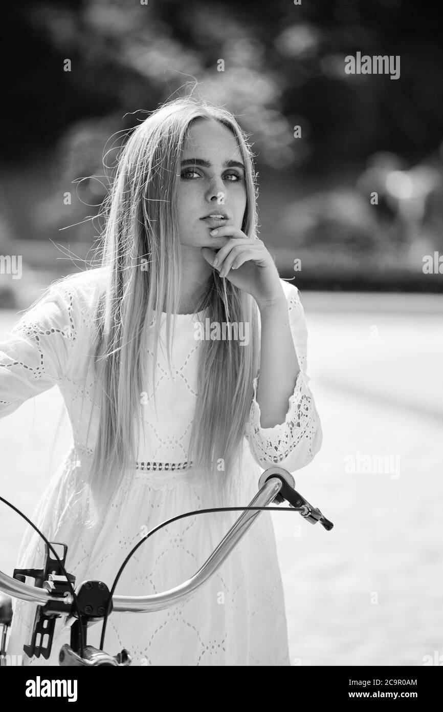 beautiful girl in white dress and blond hair with bike in sunny park looking at camera in sun light, monochrome Stock Photo