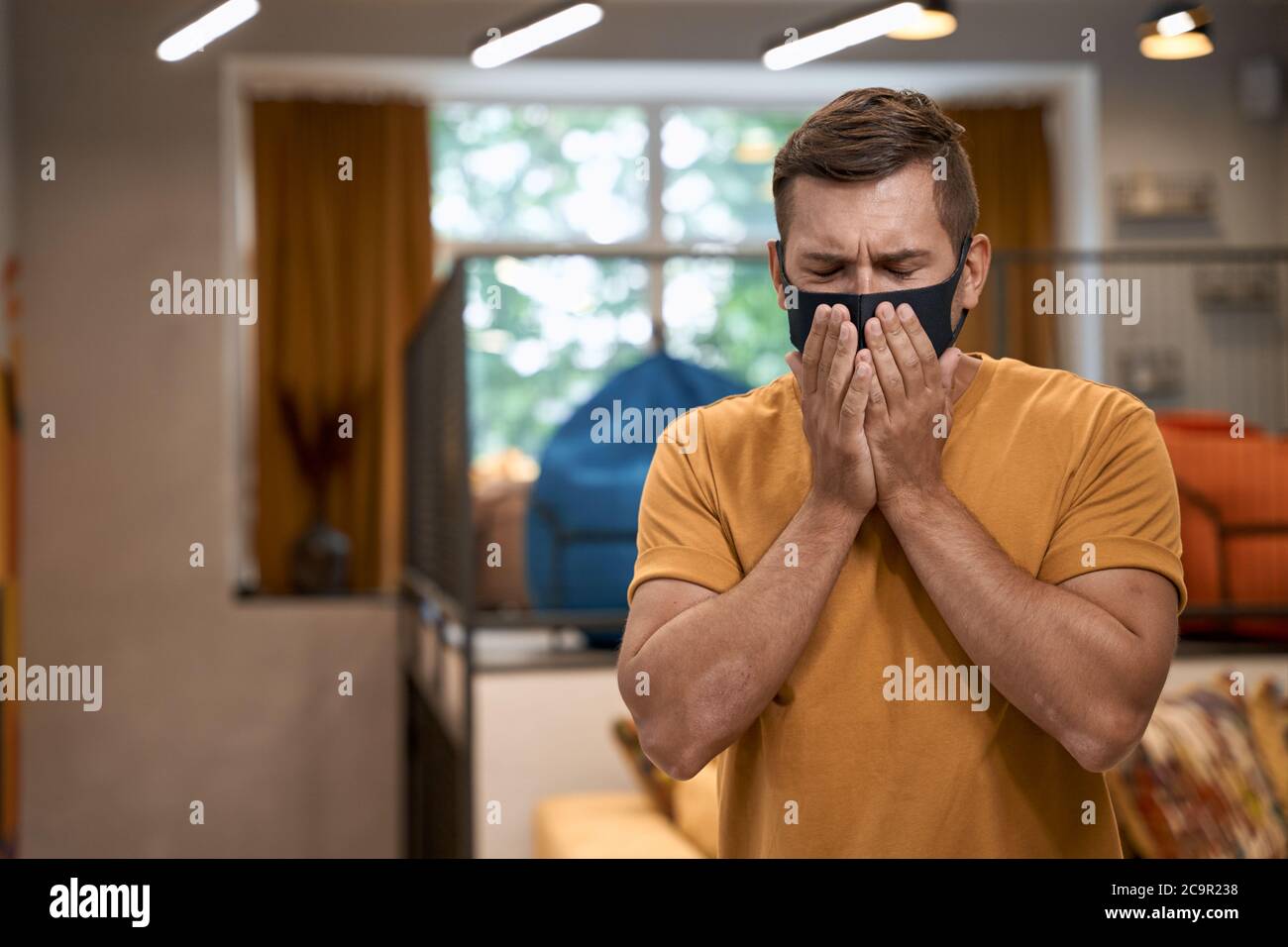 Business and Covid 19. Young man, male office worker wearing black protective mask standing in the modern office and sneezing. Social Distancing at Stock Photo
