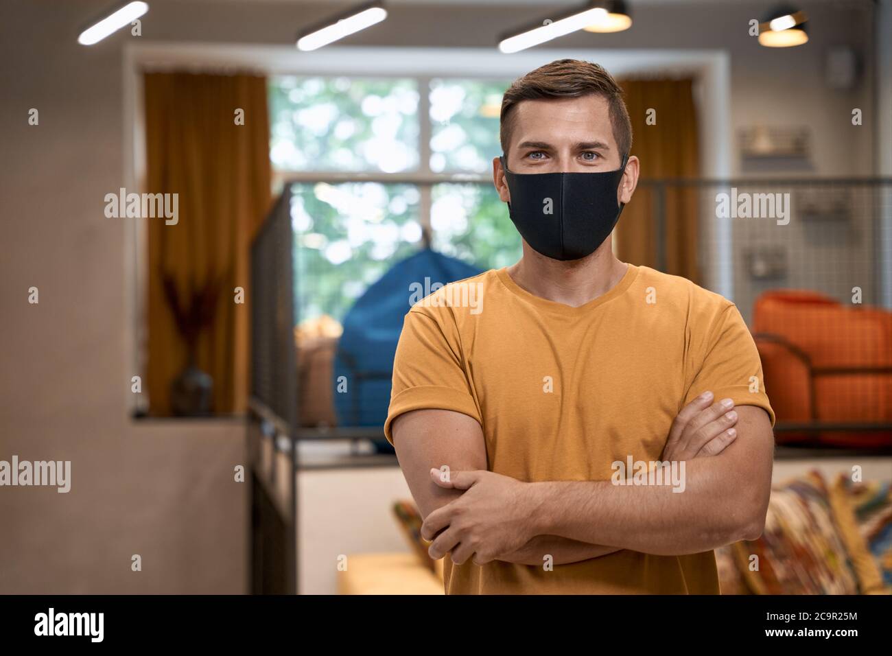 Social distancing at work. Portrait of young confident man, male office worker in protective face mask keeping arms crossed, looking at camera Stock Photo