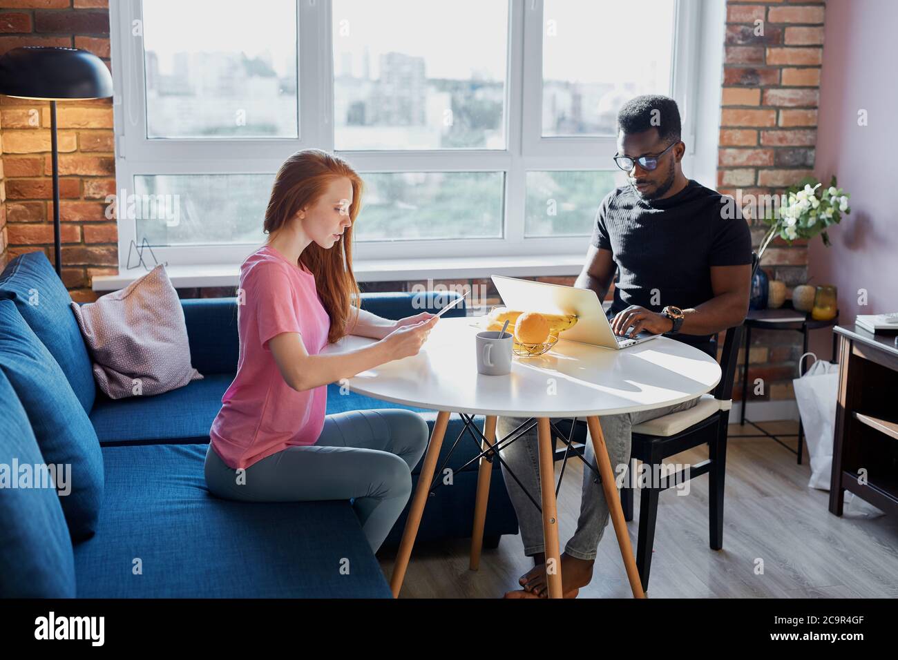 everyday life of an interracial couple, young woman sit looking at paper while black guy sit working on laptop, freelance during quarantine Stock Photo