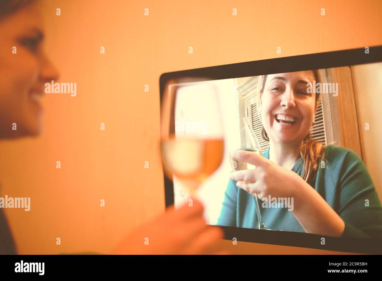 Content female friends with wineglasses communicating via video call and drinking alcohol beverages together during coronavirus epidemic Stock Photo