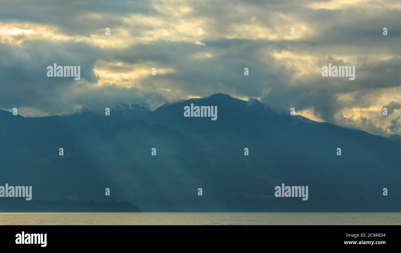 A beautiful scenery of a lake in Vancouver surrounded by mountains as the sun rays are passing through the clouds touching the crystal water Stock Photo