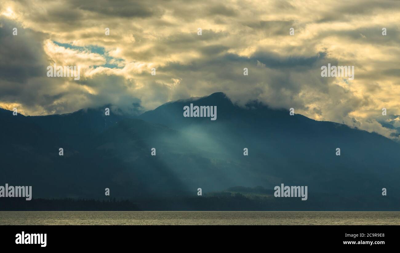 A beautiful scenery of a lake in Vancouver surrounded by mountains as the sun rays are passing through the clouds touching the crystal water Stock Photo