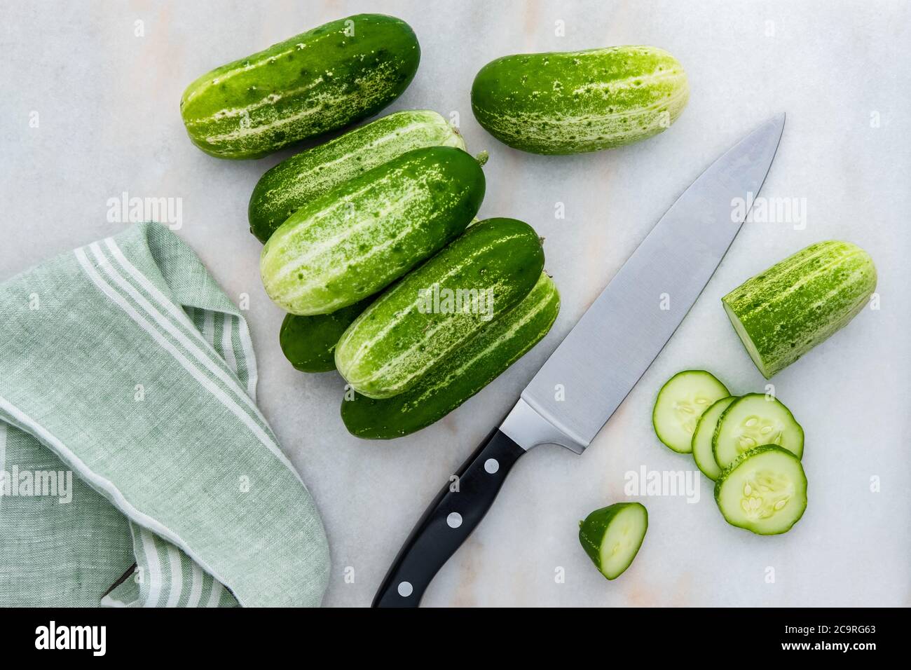 Photograph of freshly picked organic pickling cucumbers, being prepared for slicing with a chef's knife and dishtowel on kitchen counter Stock Photo