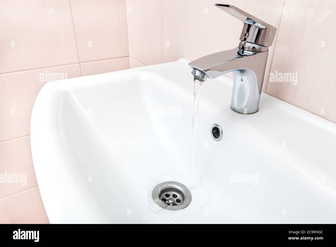 open chrome faucet water flow in the ceramic wash basin in the bathroom with a beige tile on wall. Stock Photo