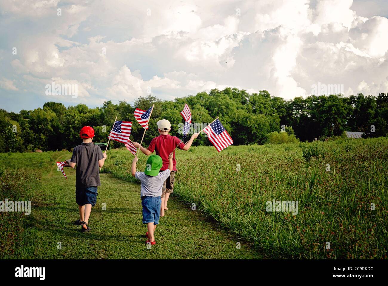 Three boys holding American Flags while walking on a country path in a field of green waving the flags Stock Photo