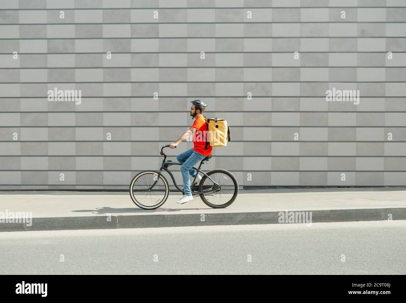 Delivery man on bicycle. Young guy with beard and big backpack rides along path in city Stock Photo