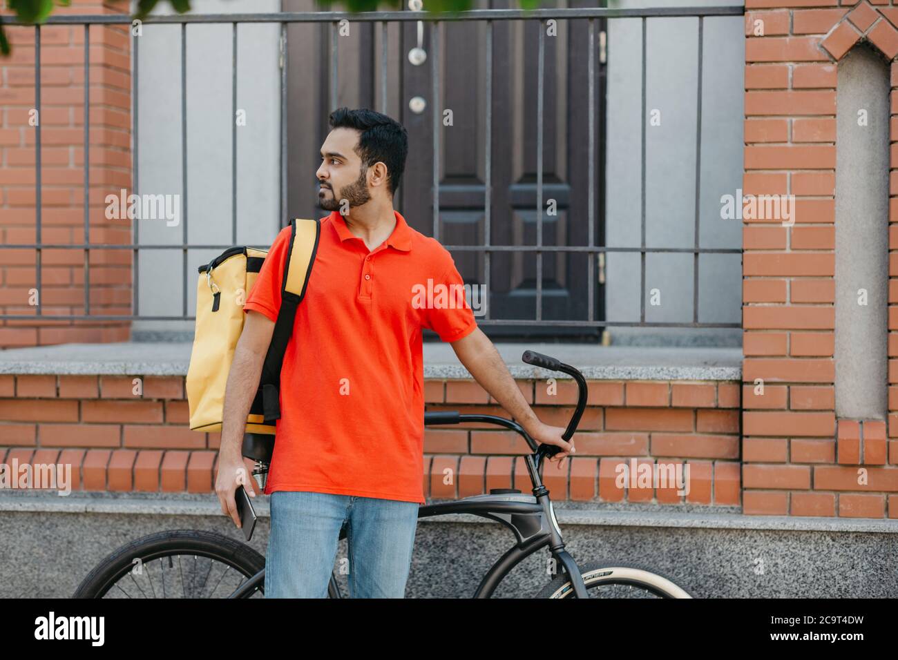 Work in city. Serious deliveryman with beard with backpack and bicycle, holding smartphone and looking for address Stock Photo