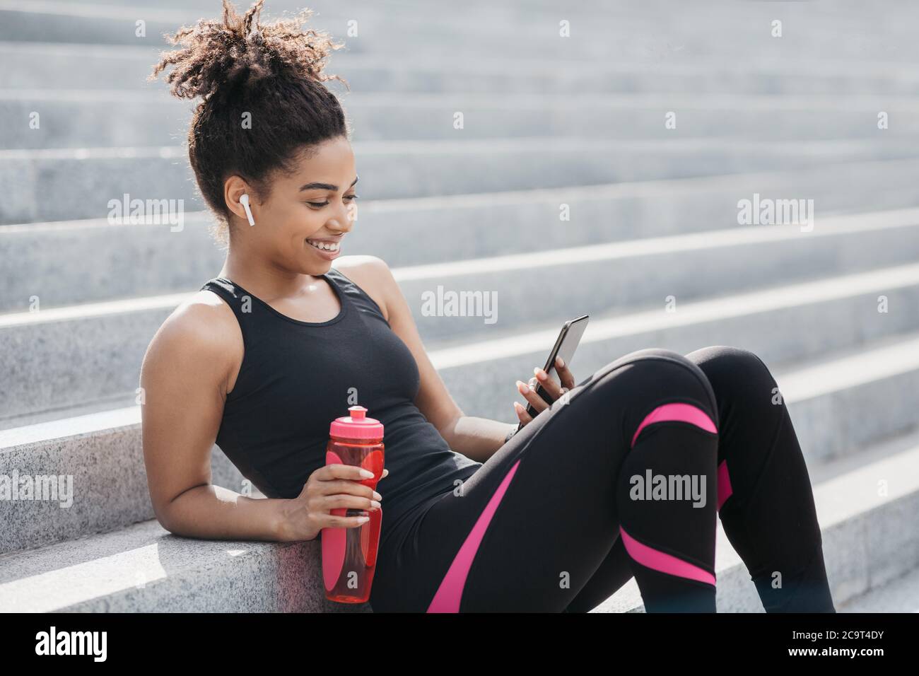 Smiling african american girl in sportswear with wireless headphones and water bottle looks at smartphone and sits on stairs Stock Photo