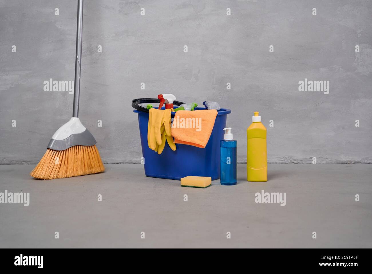 Cleaning supplies. Broom and plastic bucket or basket with cleaning products, bottles with detergents standing on the floor against grey wall. Housework, cleaning, housekeeping concept. Disinfection Stock Photo