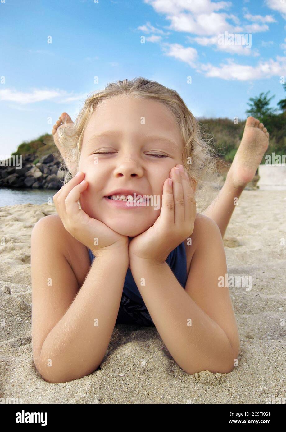 Beautiful little girl is lying on a sandy beach at stomach and laughing with close eyes Stock