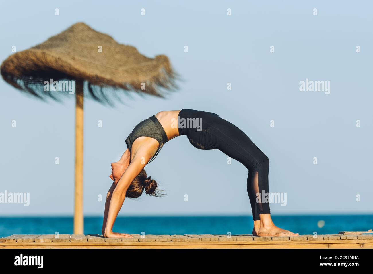 young woman doing yoga bridge exercise on the beach in front of the sea next to umbrella on a sunny day Stock Photo