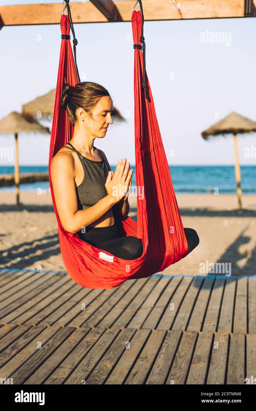 young woman doing yoga on a red swing on wooden platform by the sea on a sunny day Stock Photo