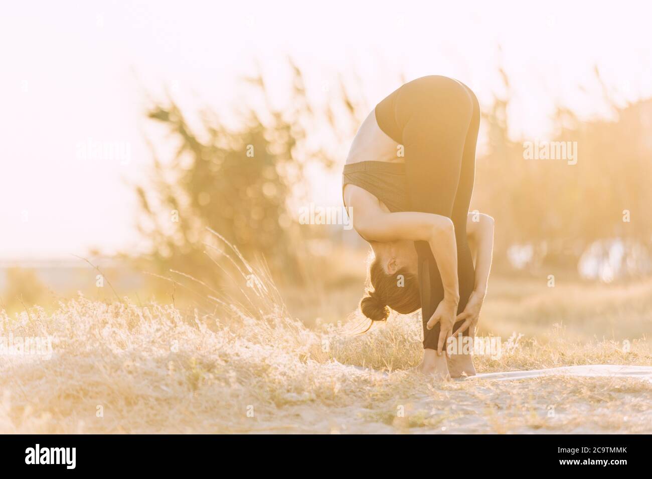 young woman doing stretching yoga exercise at golden sunset outdoors next to greenery Stock Photo