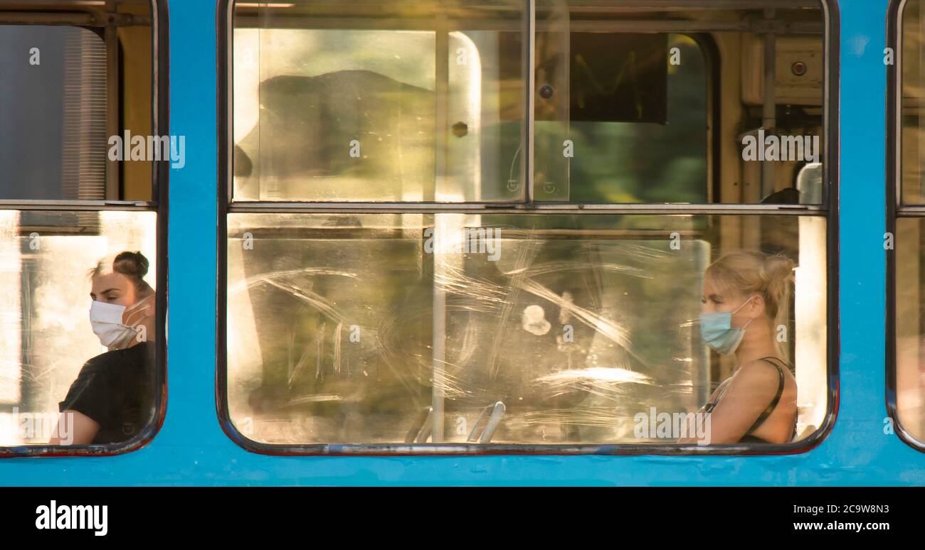 Belgrade, Serbia - July 21, 2020: Two young women wearing face surgical masks riding on a distant window seats of a tram , in summer sunset with refle Stock Photo