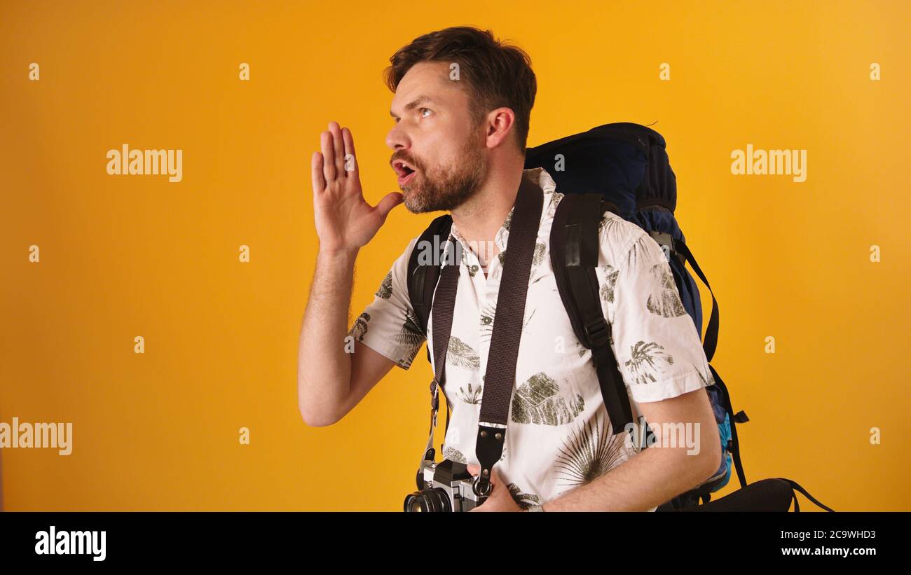 Young man traveler with backpack speaking loud with hands on the side of his face isolated on the orange background. High quality photo Stock Photo