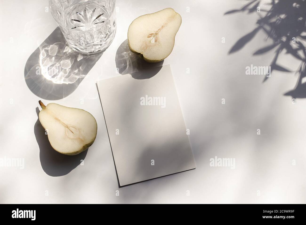 Summer stationery still life scene. Glass of water and cut pear fruit in sunlight. White table. Blank paper card, invitation mockup scene, olive Stock Photo