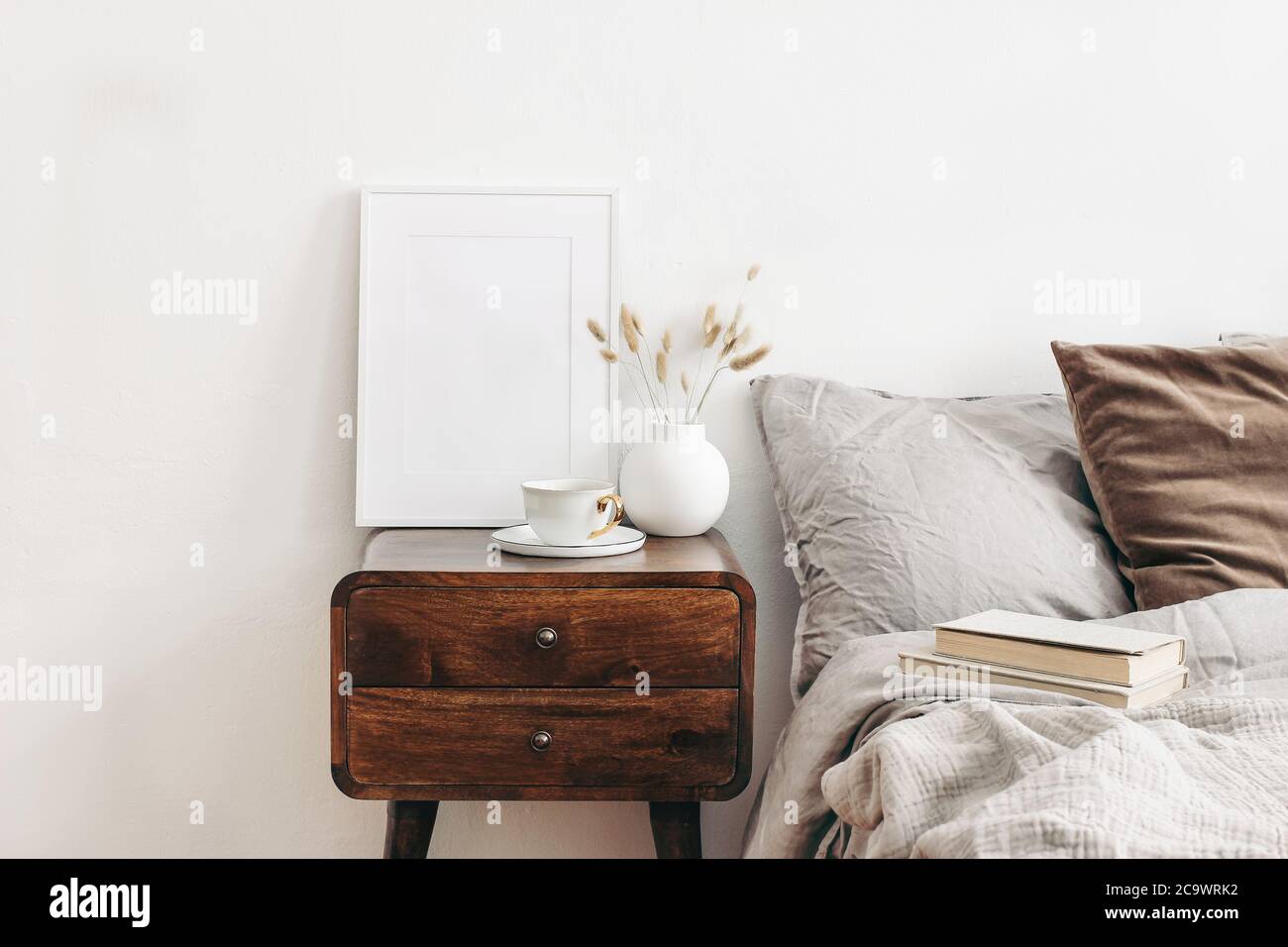 Portrait white frame mockup on retro wooden bedside table. Modern white ceramic vase with dry Lagurus ovatus grass and cup of coffee. Beige linen and Stock Photo