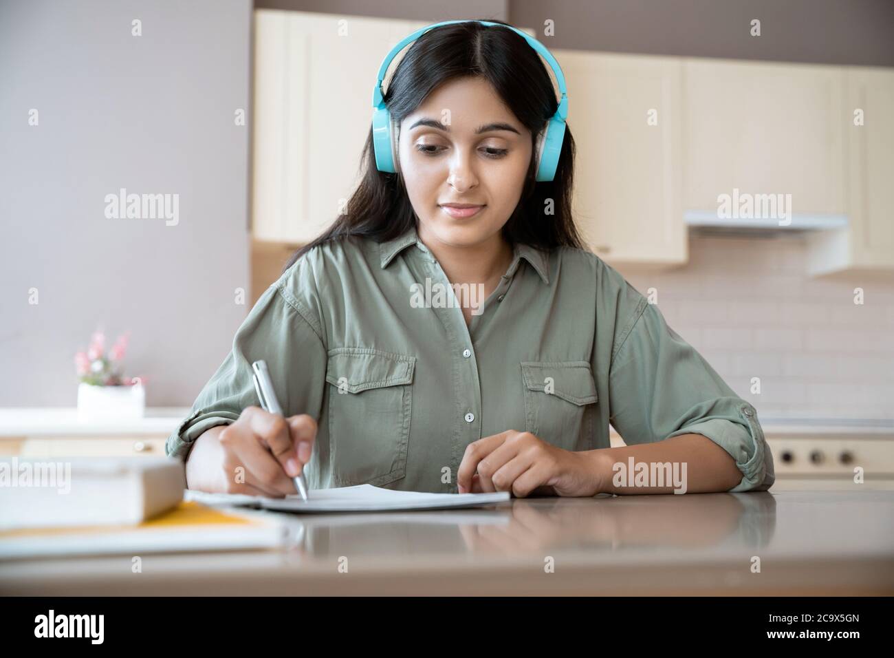 Indian girl wear headphones writing in notebook listening audio lesson at home. Stock Photo