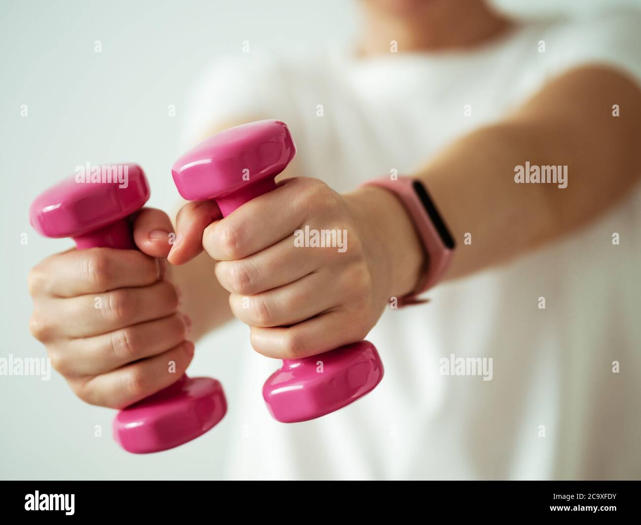 Unrecognizable young woman in white shirt with pink wearable device and bright pink colored dumb-bell in hands. Focus on barbells in female hands Stock Photo