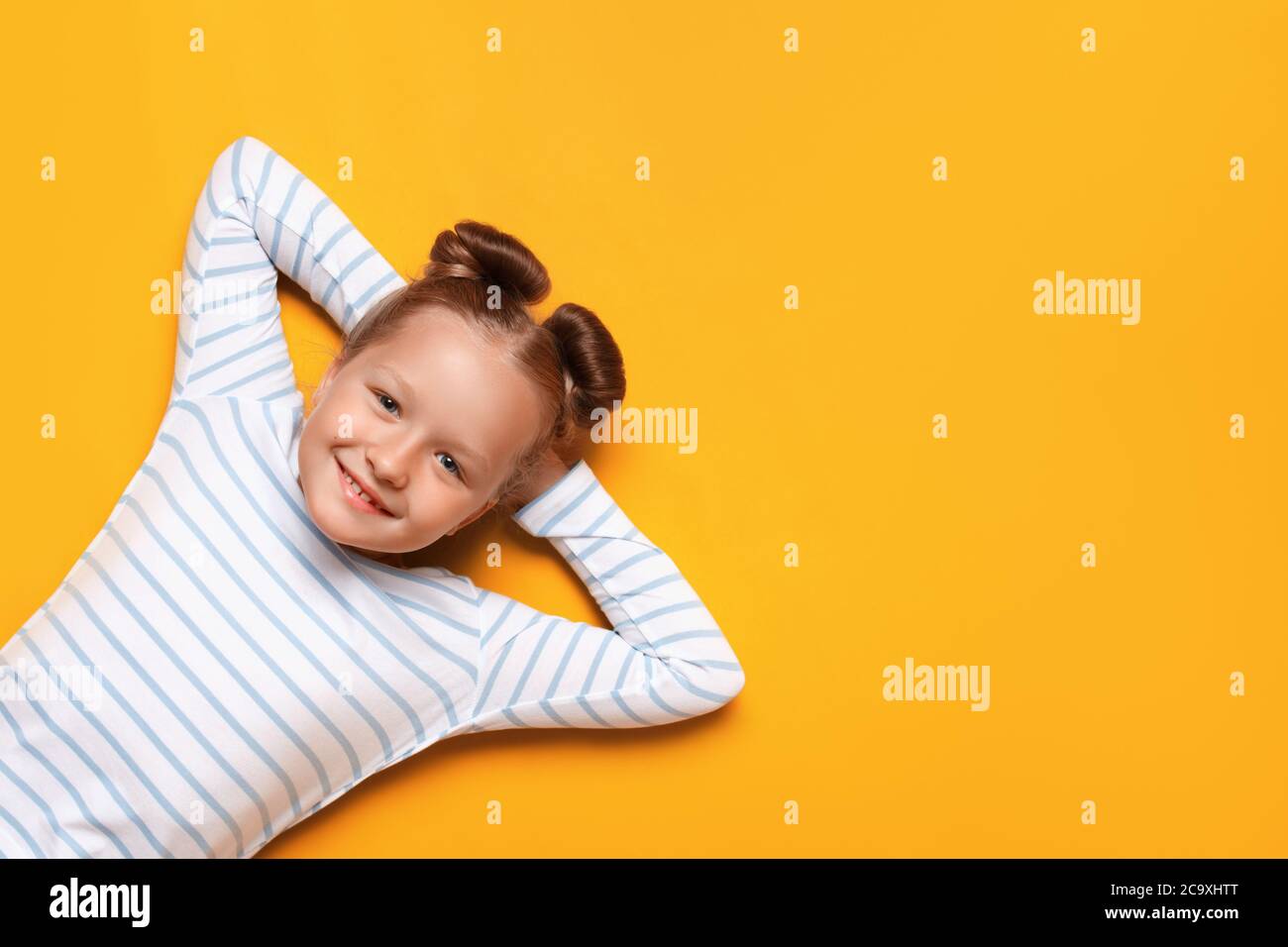 Portrait of a cute attractive adorable little girl with hair buns on a yellow background. The child lies with his hands folded behind his head. Close- Stock Photo