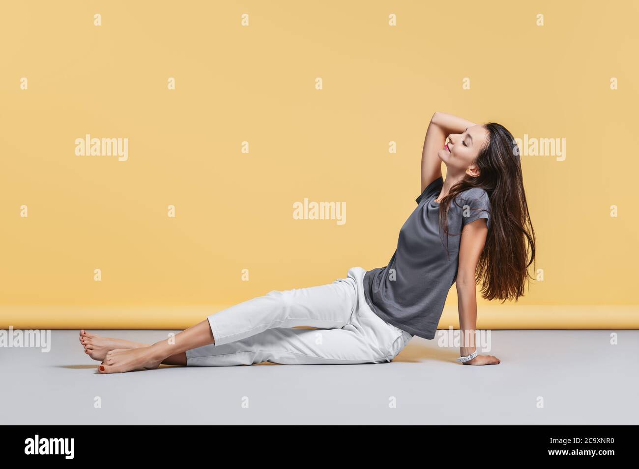 Happy barefoot young woman in casual trousers and shirt sits on the floor in studio on yellow background Stock Photo