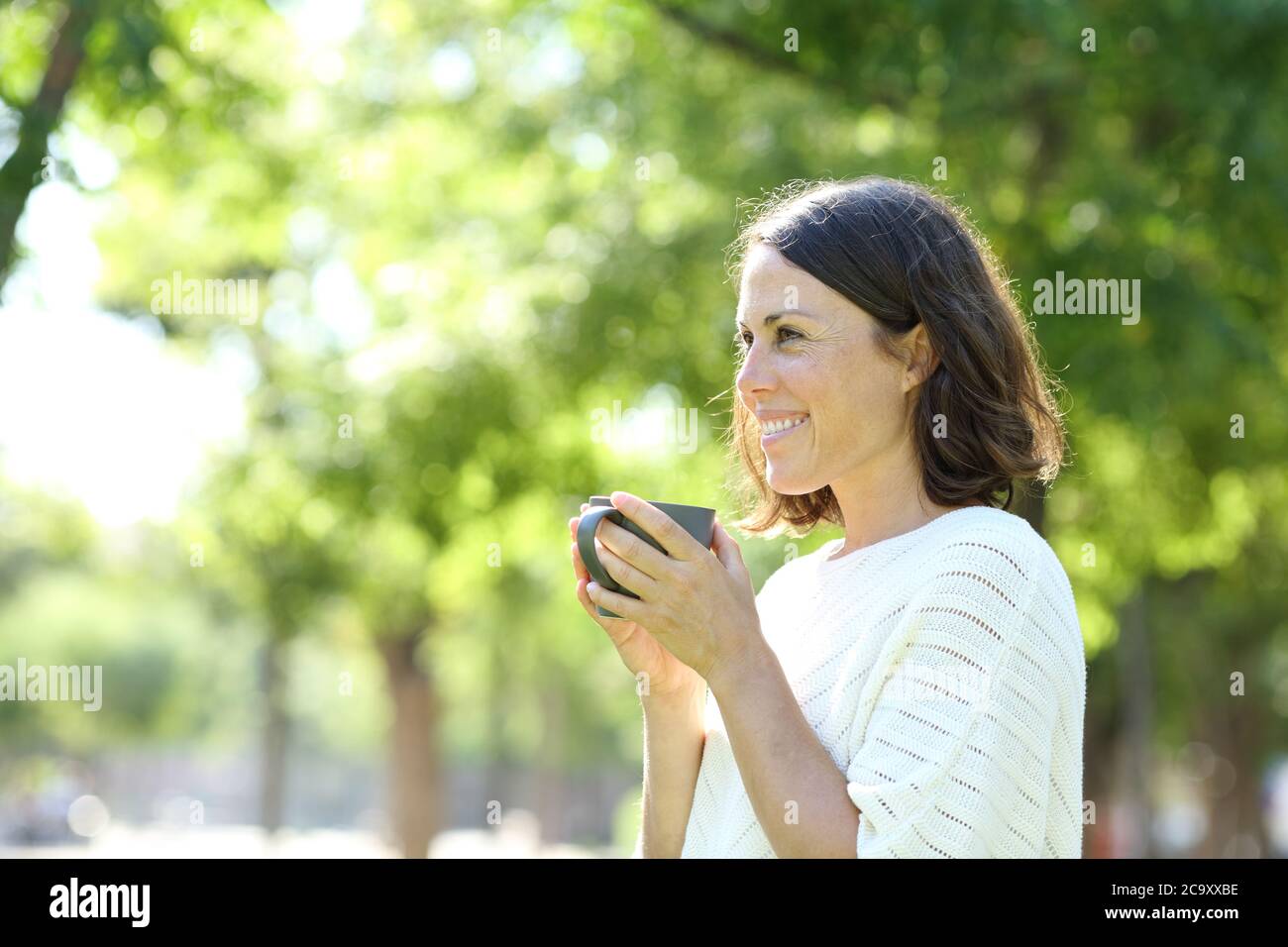 Happy adult woman holding a cup of coffee standing in a green park in ...
