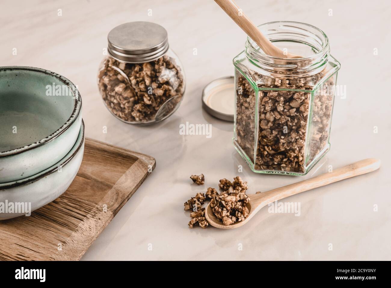 Homemade healthy and nutritious keto diet breakfast granola in glass jars with wooden spoons and blue ceramic bowls on marble kitchen table Stock Photo