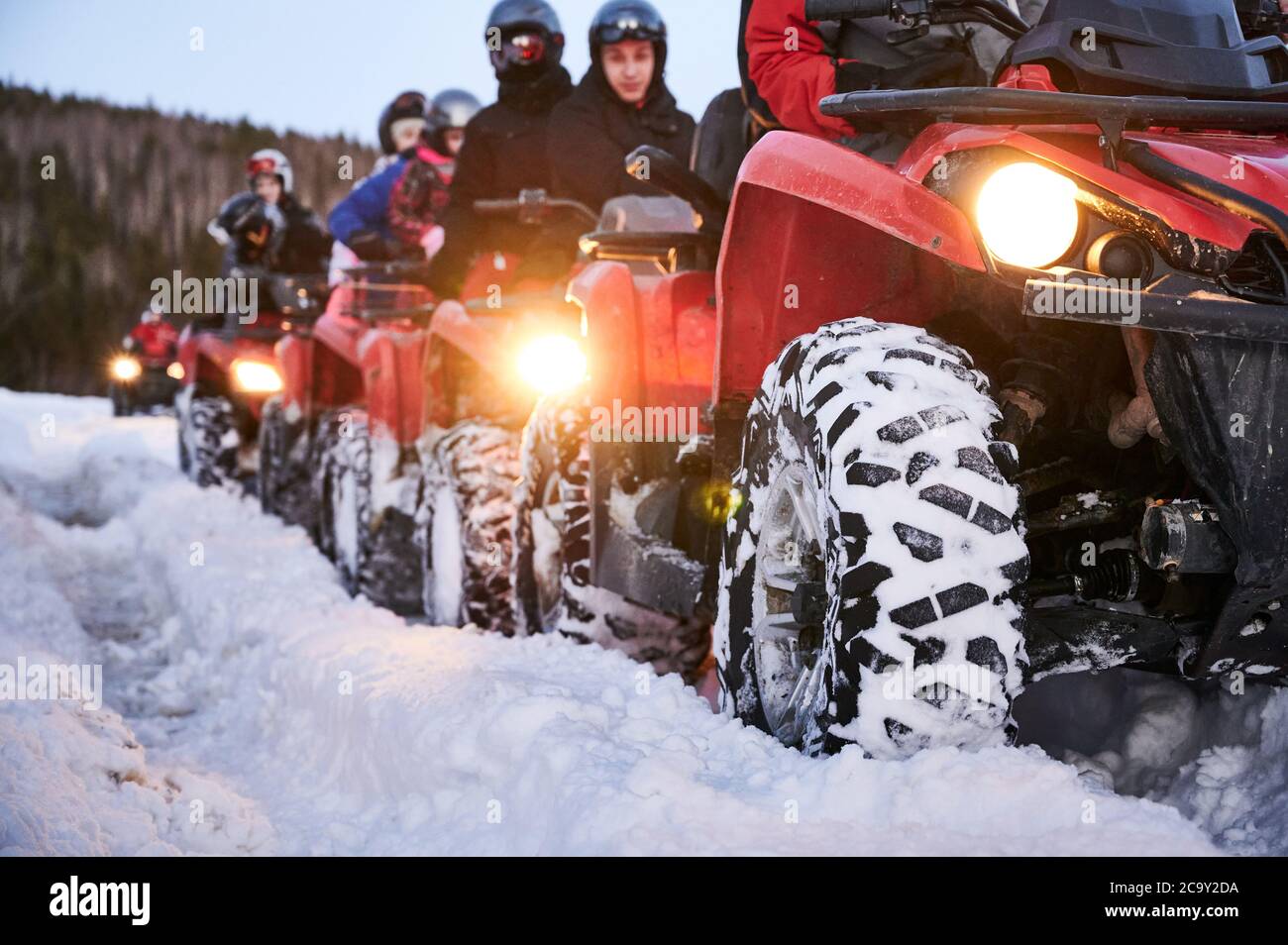 Focus on the wheel. People riding red all-terrain vehicles down snowy hill. Quad riders driving quad bikes with black snowy wheels on snow-covered trail. Concept of winter activities and quad biking. Stock Photo