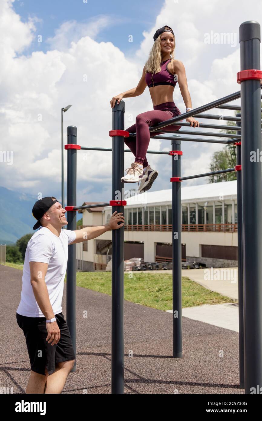 Outdoor training - young caucasian couple exercising on horizontal bar near fitness centre. Male Instructor talking with sportive girl on bar. Stock Photo