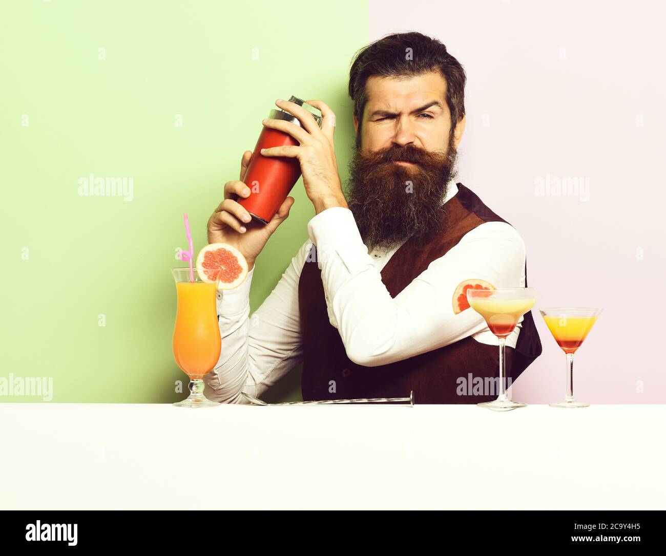 handsome bearded barman with long beard and mustache has stylish hair on funny face holding shaker and made alcoholic cocktail in vintage suede leather waistcoat, on purple green studio background Stock Photo