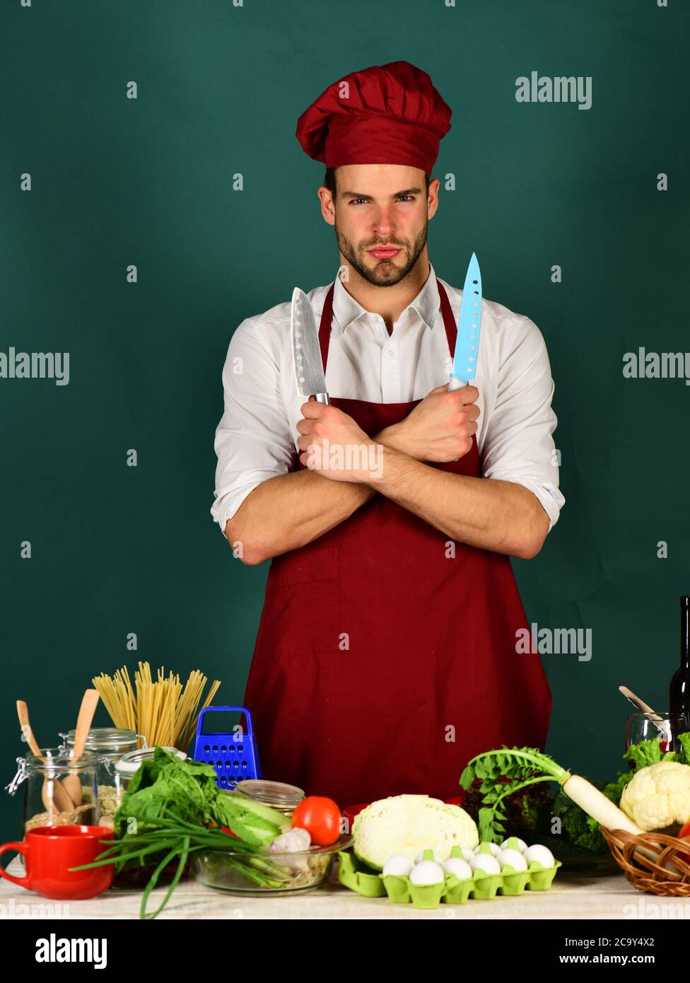 https://c8.alamy.com/comp/2C9Y4X2/man-in-cook-hat-and-apron-chef-with-confident-face-holds-blue-and-metal-knives-on-dark-green-background-cook-works-in-kitchen-near-table-with-vegeta-2C9Y4X2.jpg