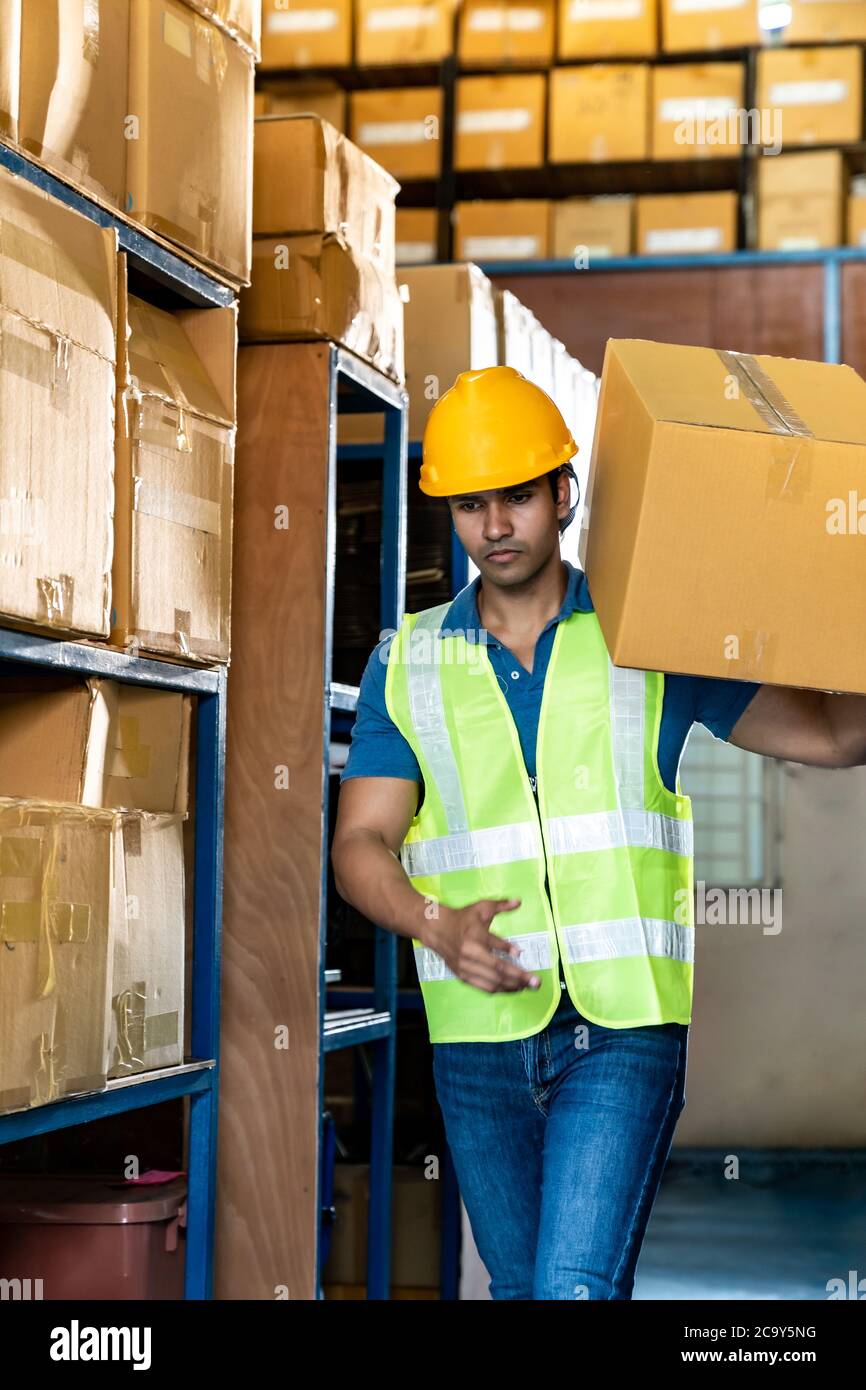 Portrait of Indian asian warehouse worker hold cardboard box packaging on his shoulder in warehouse distribution center environment. Using in business Stock Photo