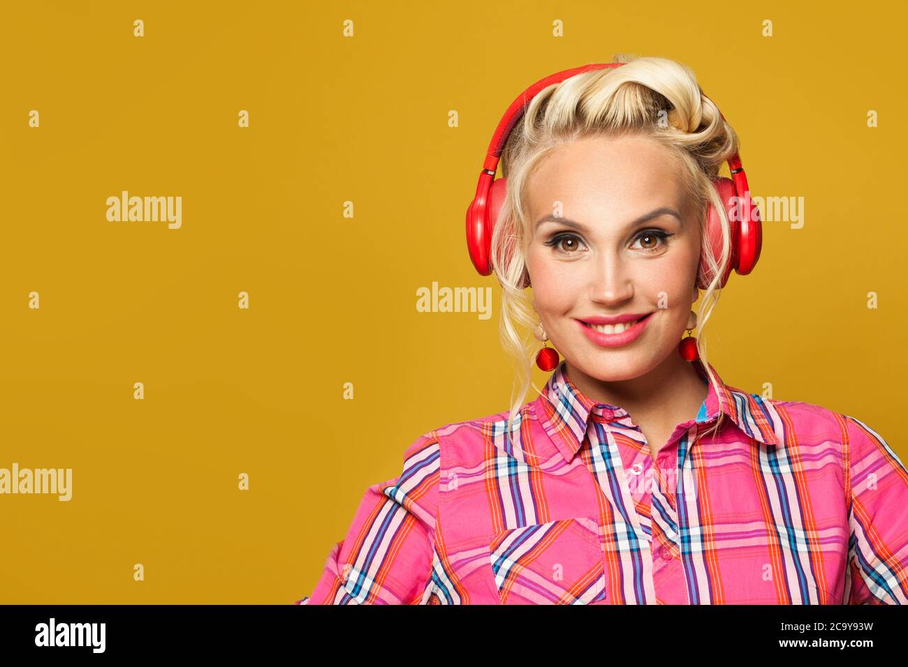 Pretty pinup girl listening music with headphones on bright yellow background. Pretty retro model woman with red lips makeup and vintage fashion hairs Stock Photo