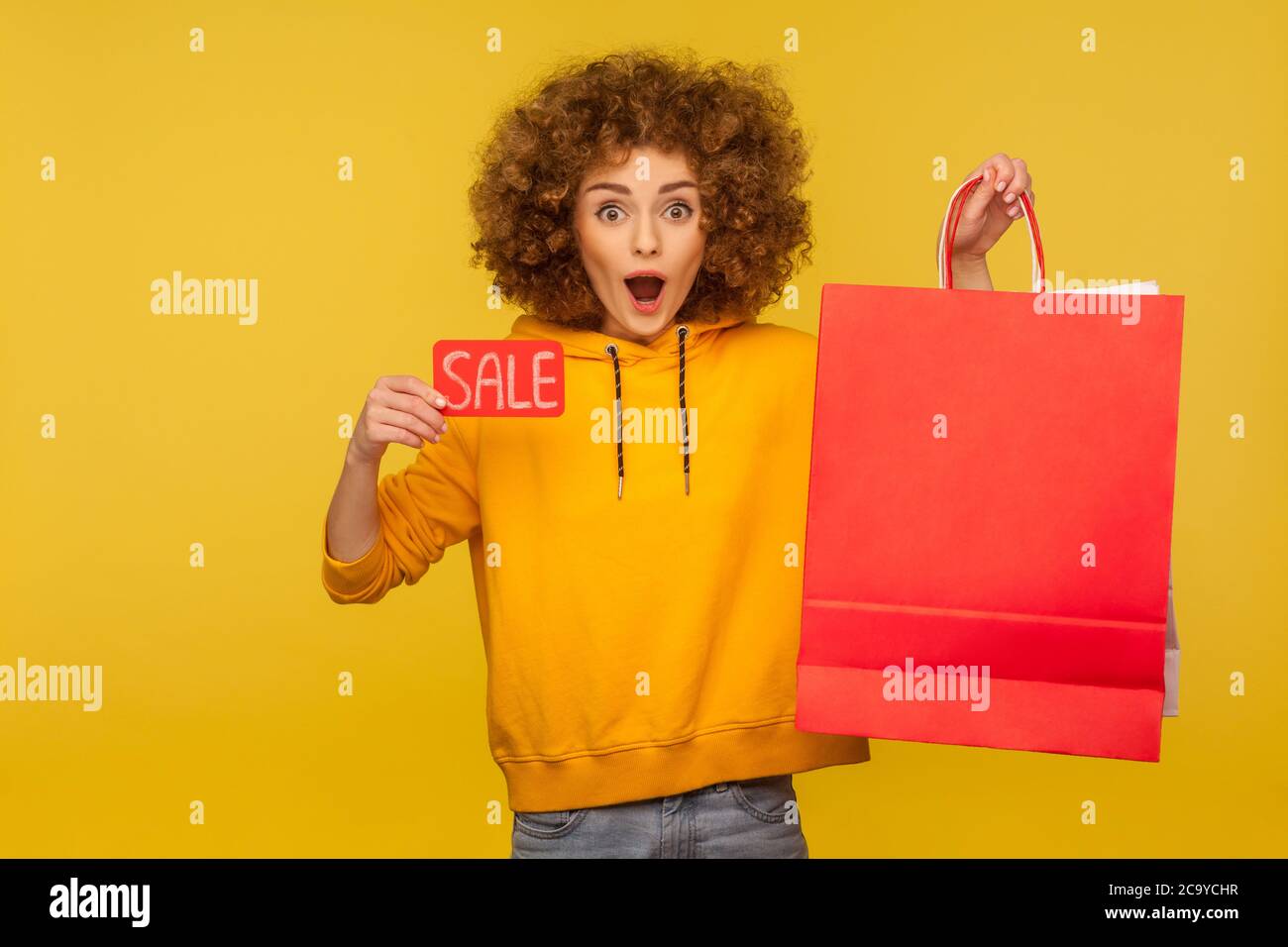 Portrait of shocked surprised curly-haired hipster girl in hoodie holding shopping bags with blank area for advertising text and showing Sale inscript Stock Photo