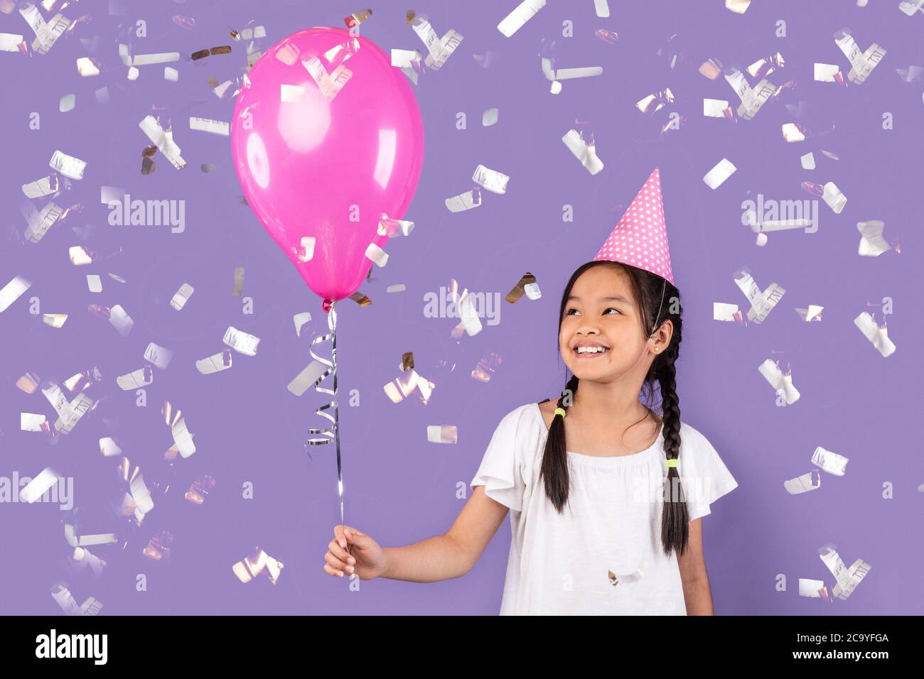 Girl Holding Balloon Celebrating Birthday Under Falling Confetti, Purple Background Stock Photo